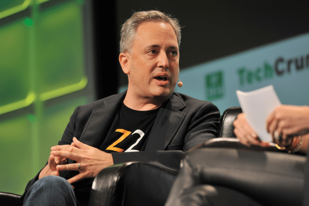 A man with graying hair wearing a dark jacket and a black t -shirt with orange lettering is speaking, seated in a black leather chair at an event with a green and white TechCrunch logo in the background.