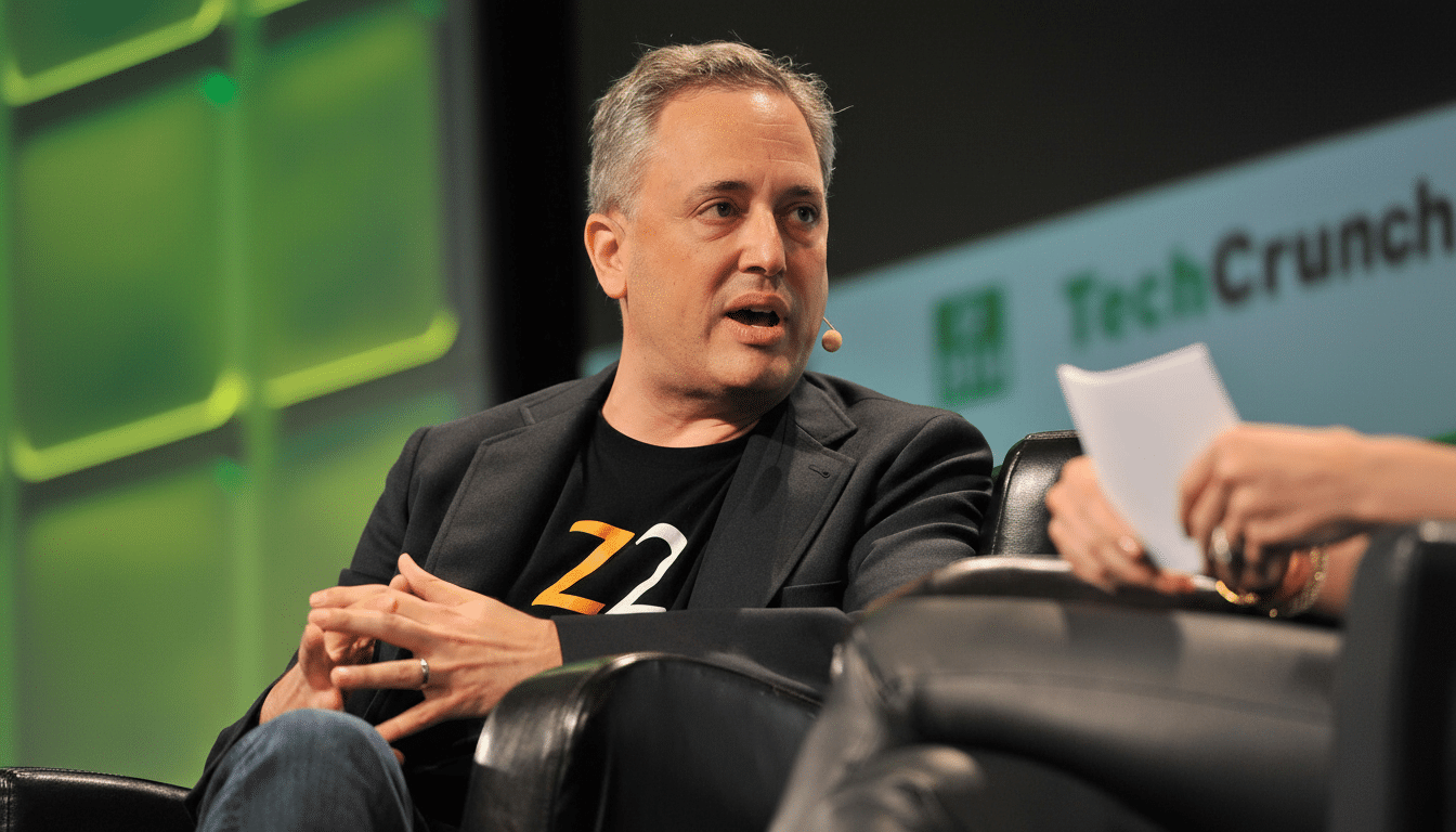 A man with graying hair wearing a dark jacket and a black t -shirt with orange lettering is speaking, seated in a black leather chair at an event with a green and white TechCrunch logo in the background.