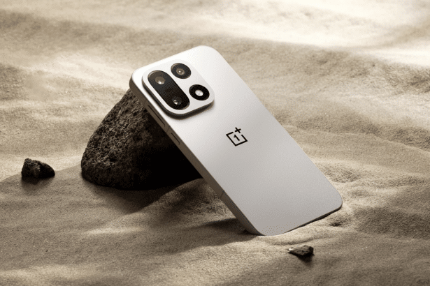 A silver smartphone with a black camera module leaning against a dark rock on a textured sandy surface with small pebbles scattered around. Filename : silver smartphone sand rock.png