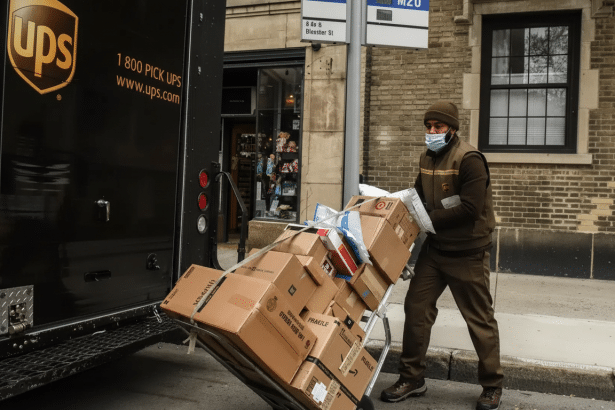 A UPS delivery driver wearing a face mask pushes a hand truck loaded with packages next to a brown UPS truck on a city street.
