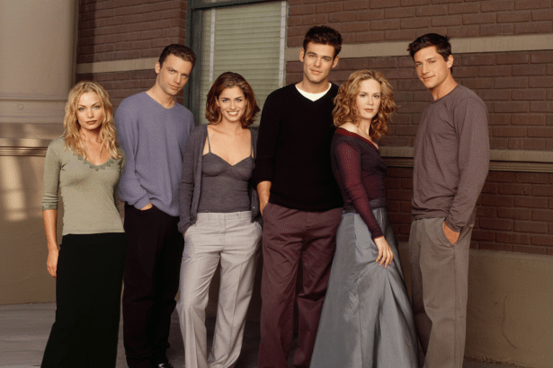 Six young adults, three men and three women , stand in a line against a brick building background. They are dressed in casual clothing .