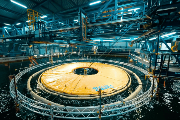 An indoor facility with a large water tank , showing a circular yellow floating device labeled SIZABLE ENERGY surrounded by a metal frame structure , with industrial walkways and equipment in the background.