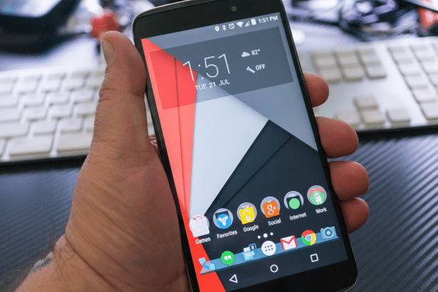 A hand holding a smartphone displaying the home screen with various app icons, time, and weather, set against a blurred background of a white keyboard