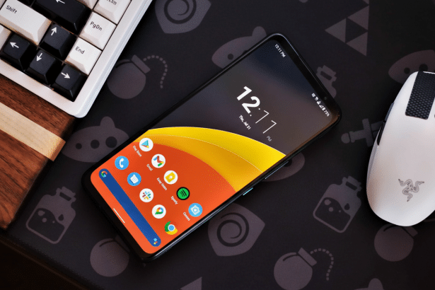 A smartphone displaying the time 1 2: 11 PM on a black mat with a white keyboard and mouse.