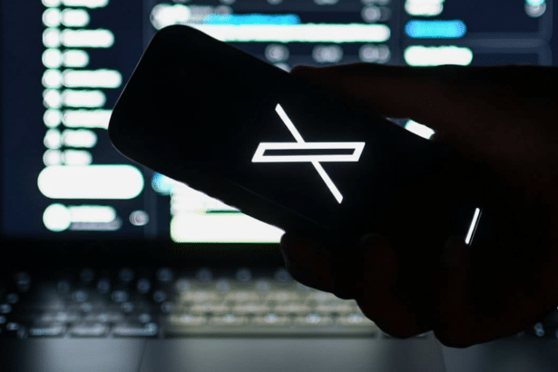 A hand holding a smartphone displaying the X logo, with a blurred background of a laptop screen showing lines of code.