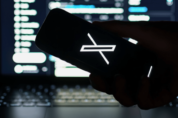 A hand holds a smartphone displaying the X logo, sil houetted against a laptop screen showing code.