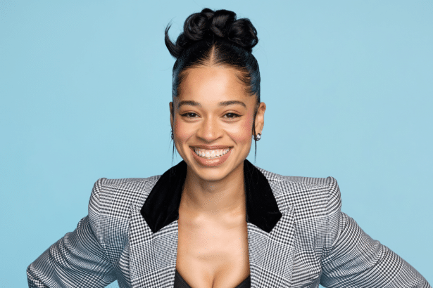 A smiling woman with dark hair in an updo and a black and white plaid blazer, set against a light blue background, cropped to a 1 6:9 aspect ratio. Filename : smiling womanpl aidblazer 16 9.png