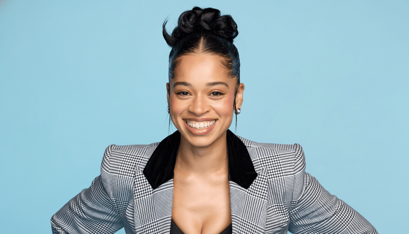 A smiling woman with dark hair in an updo and a black and white plaid blazer, set against a light blue background, cropped to a 1 6:9 aspect ratio. Filename : smiling womanpl aidblazer 16 9.png