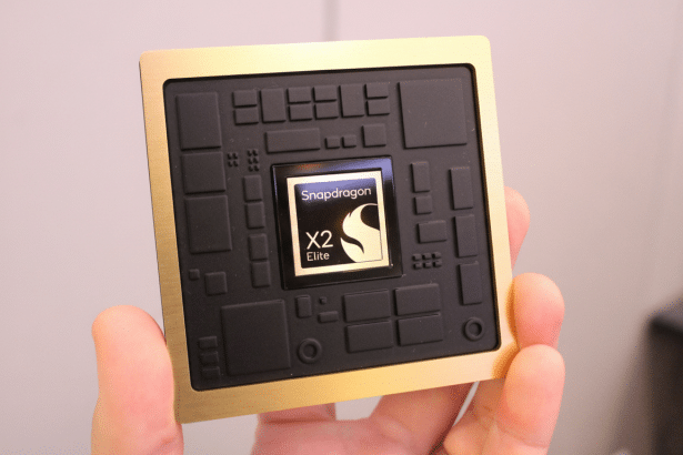 A hand holds a gold and black Snapdragon X2 Elite chip, with the logo visible on the central square. The background is a slightly blurred office- like setting.