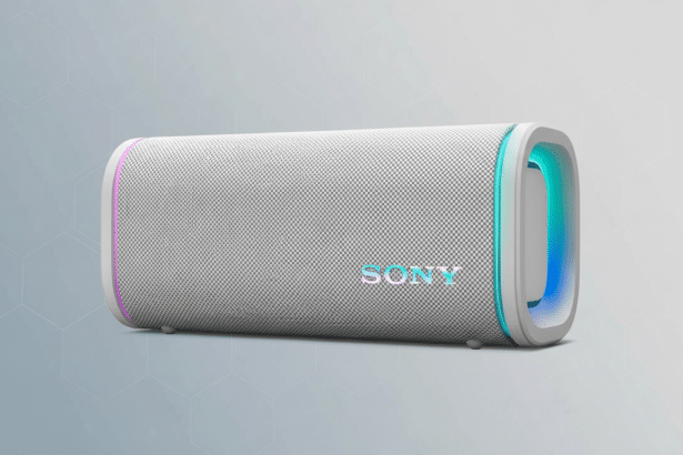 A white Sony portable Bluetooth speaker with colorful LED lights on the ends and the SON Y logo illuminated in the center, set against a professional gray background with a subtle geometric pattern. Filename : sonybluetooth speakerprofessional .png