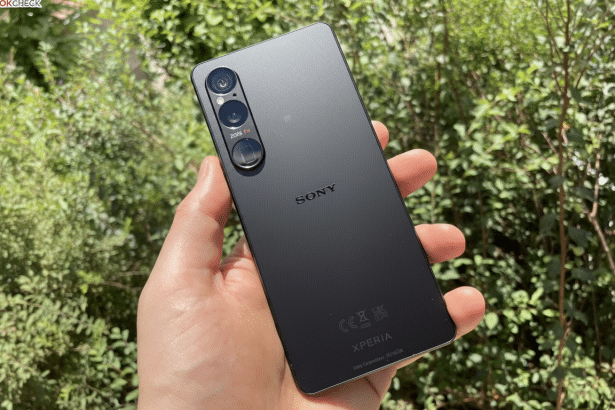 A hand holding a black Sony Xperia smartphone with a triple camera setup on the back, against a blurred green foliage background. Filename : sony xperia hand held 1 6x 9.png