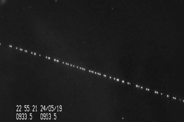 A black and white image showing a diagonal line of bright dots , likely satellites , against a dark sky with faint stars. Time and date information are visible in the bottom left corner .