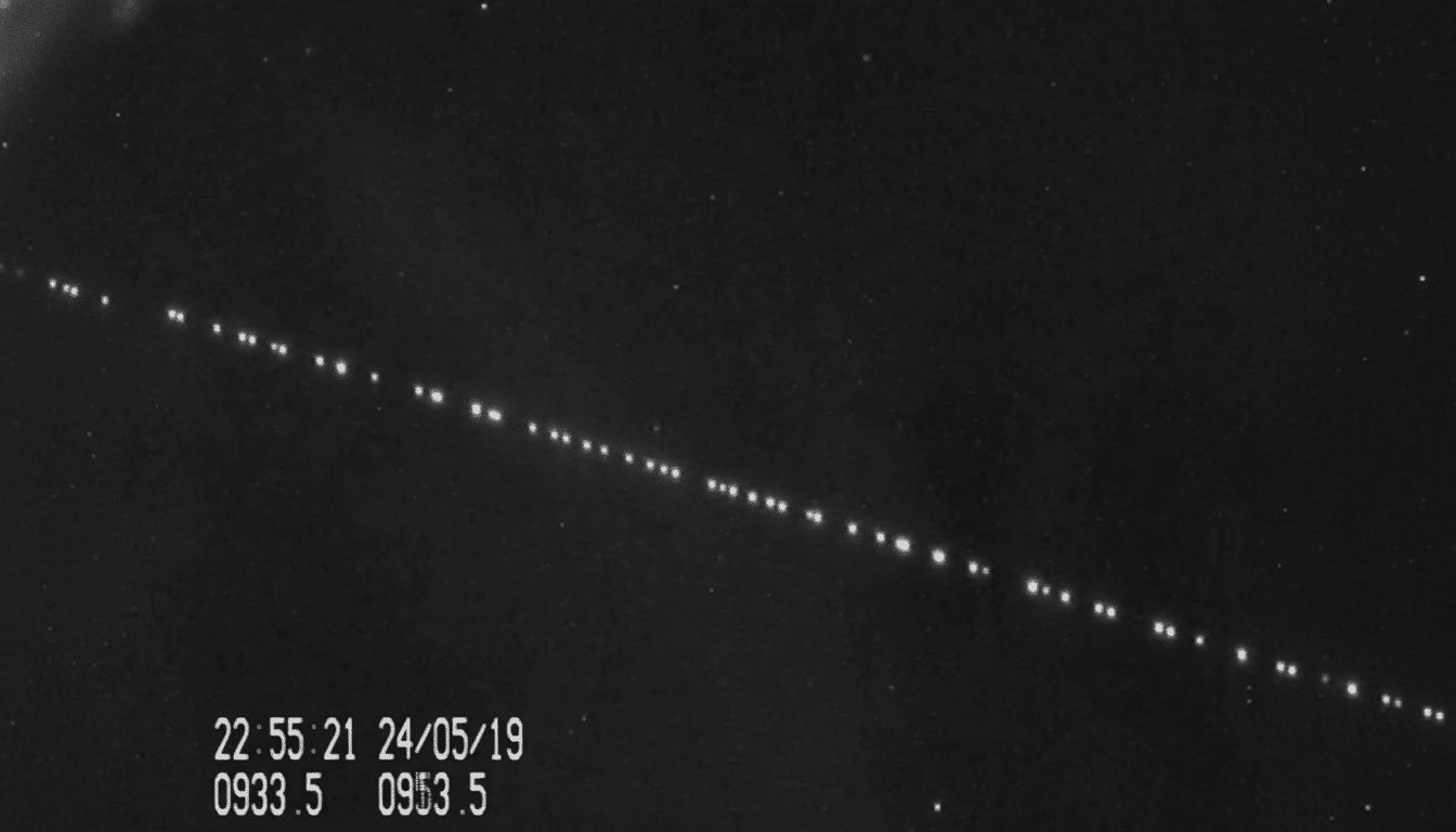 A black and white image showing a diagonal line of bright dots , likely satellites , against a dark sky with faint stars. Time and date information are visible in the bottom left corner .
