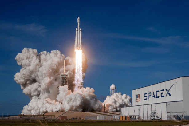 A SpaceX Falcon Heavy rocket launching from a launchpad , emitting a large plume of smoke and fire . The SpaceX logo is visible on a building to the right.