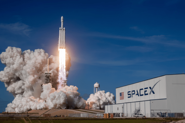A SpaceX Falcon Heavy rocket launching from a launchpad, emitting a large plume of smoke and fire against a clear blue sky, with a SpaceX building vis