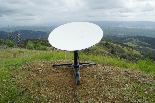 A Star link satellite dish positioned on a grassy hill with a panoramic view of rolling hills and a cloudy sky in the background.