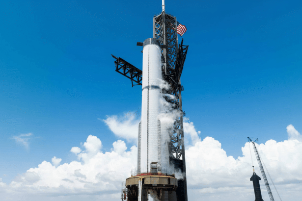 A large white rocket stands tall on a launchpad under a blue sky with white clouds, with the American flag visible near its top .