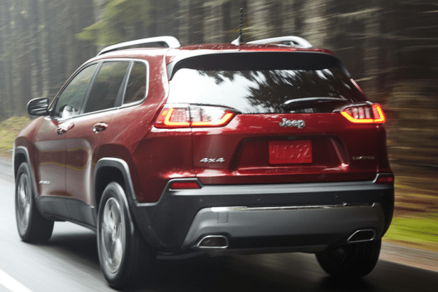 A red Jeep Cherokee driving on a paved road surrounded by trees, viewed from the rear passenger side. Filename : red jeep cherokee1 6x9. png