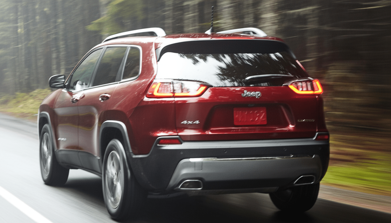 A red Jeep Cherokee driving on a paved road surrounded by trees, viewed from the rear passenger side. Filename : red jeep cherokee1 6x9. png