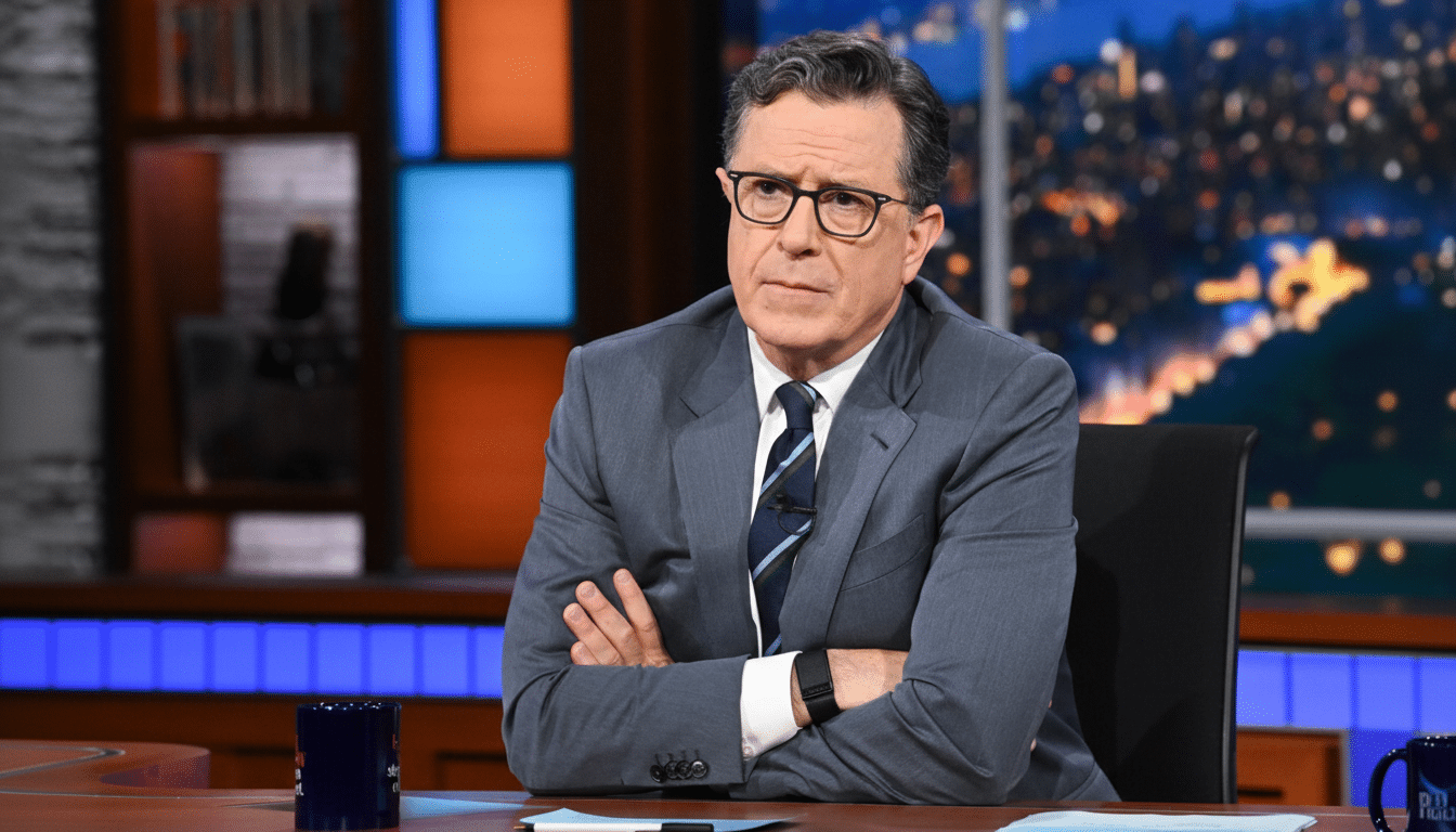A man in a suit and glasses, Stephen Colbert, sits at a desk with his arms crossed, looking thoughtfully towards the left. The background features bookshelves and a cityscape .