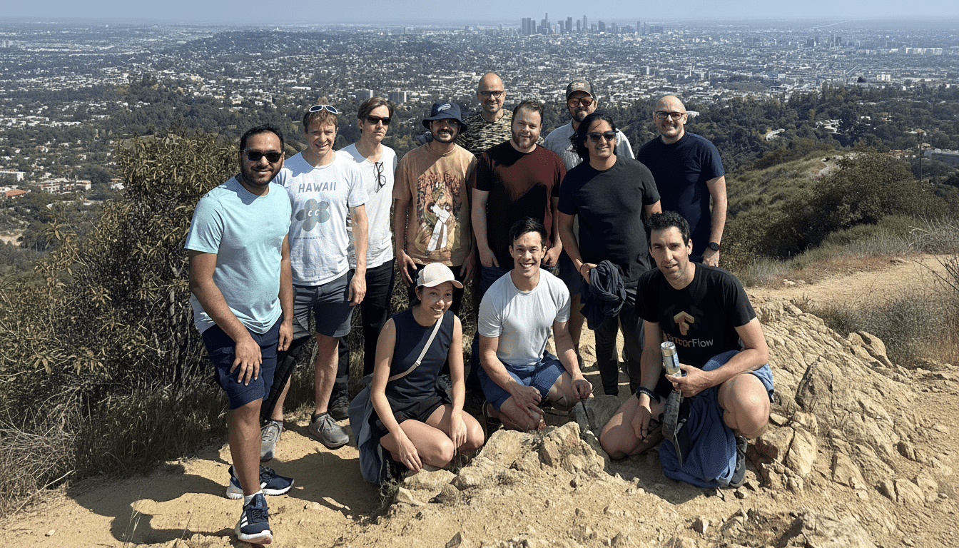 A group of thirteen people posing on a rocky trail with a vast city panorama in the background.
