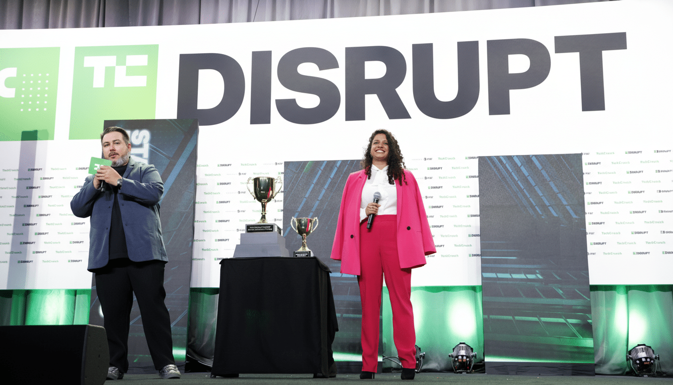 alt text: Two presenters , a man and a woman in a pink suit, stand on a stage with a TechCrunch Dis