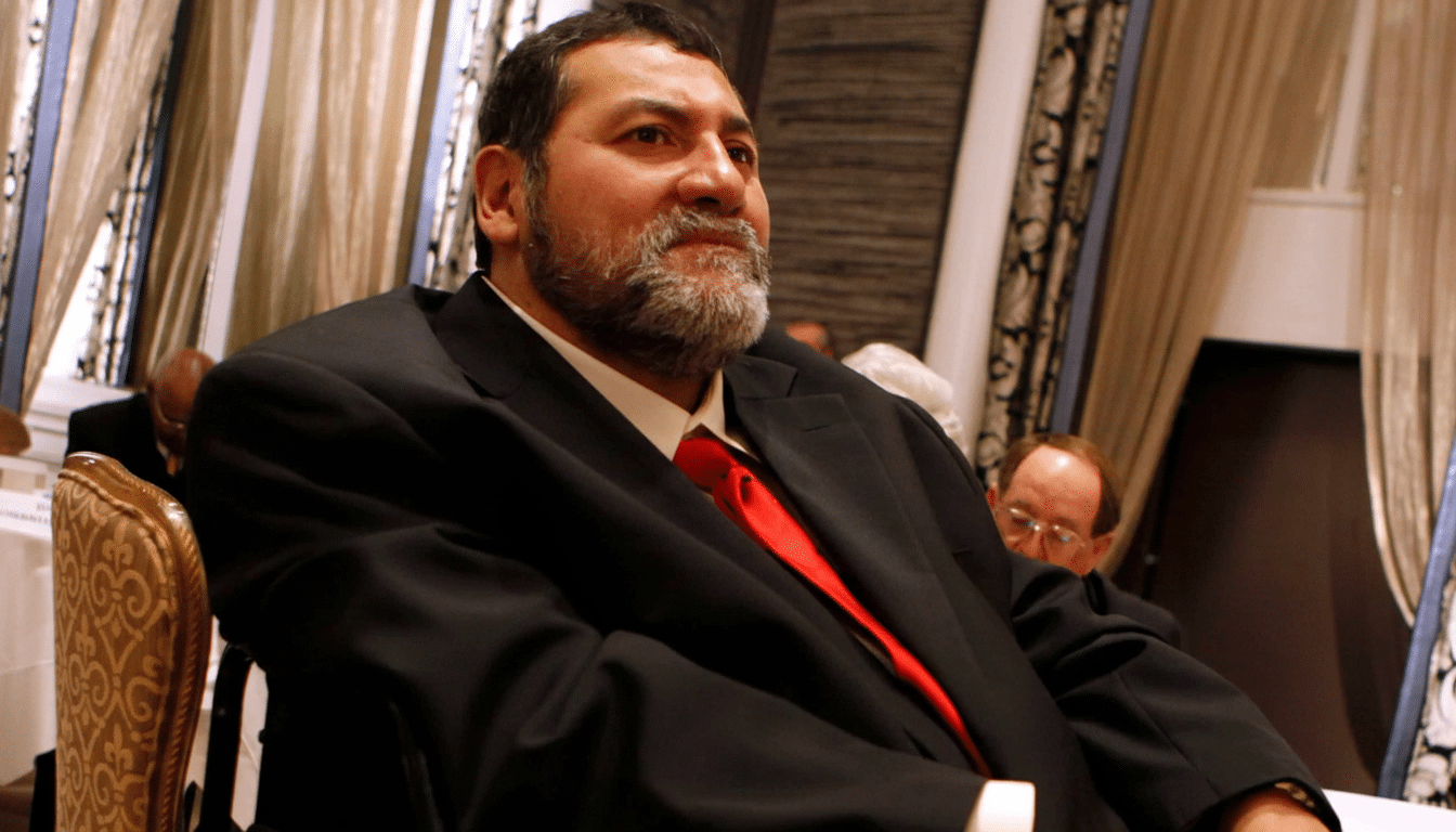 A formally dressed man with a beard, looking to the side, seated in a room with patterned curtains.