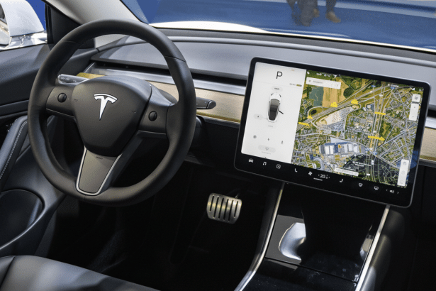 Interior shot of a Tesla vehicle , showing the steering wheel with the Tesla logo and a large central touchscreen displaying navigation and car controls. Filename : teslainterior dashboard . png