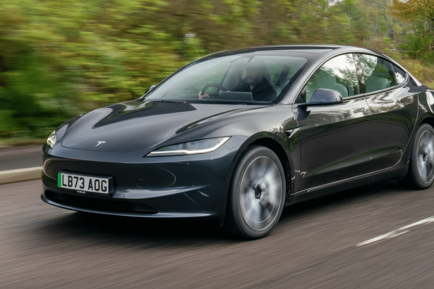 A dark gray Tesla Model 3 driving on a road with blurred green foliage in the background, captured in a 16:9 aspect ratio.