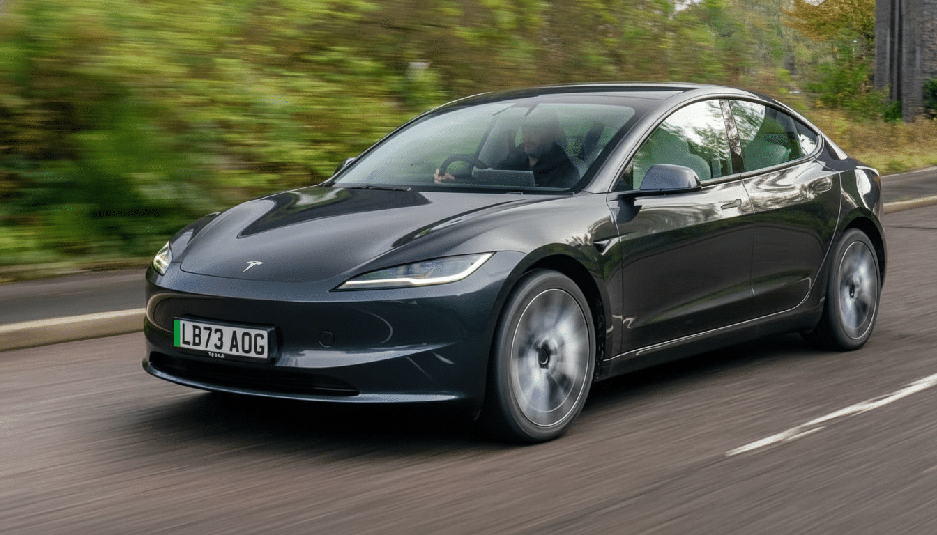 A dark gray Tesla Model 3 driving on a road with blurred green foliage in the background, captured in a 16:9 aspect ratio.