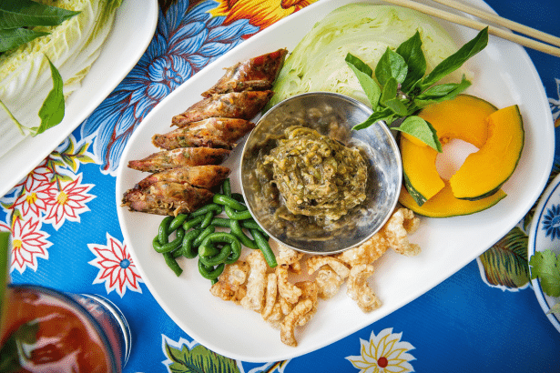 A professional overhead shot of a Thai food platter, featuring various dishes like grilled sausages, green beans, cabbage, pumpkin slices, a bowl of d
