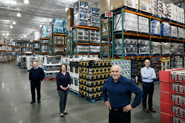 Four people standing in a large warehouse with tall shelves filled with products, resized to a 16: 9 aspect ratio.