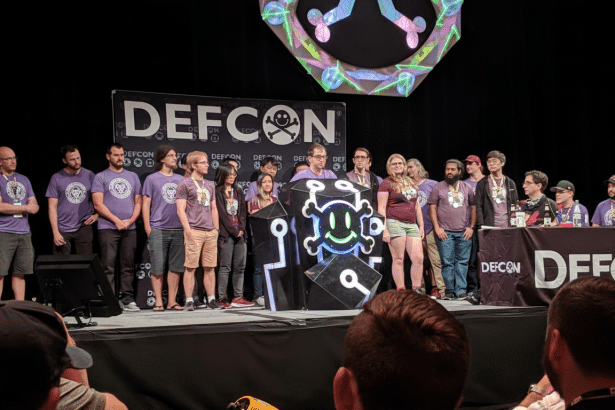 A group of people stand on a stage with a DEFCON banner in the background. A podium with a skull and crossbones logo is in the center .