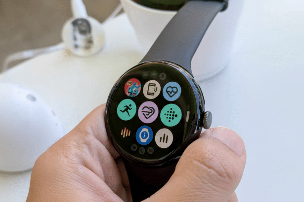 A close -up shot of a Google Pixel Watch being held, displaying various colorful app icons on its circular screen, with a blurry background showing parts of a white mouse , earbuds , and a plant pot .