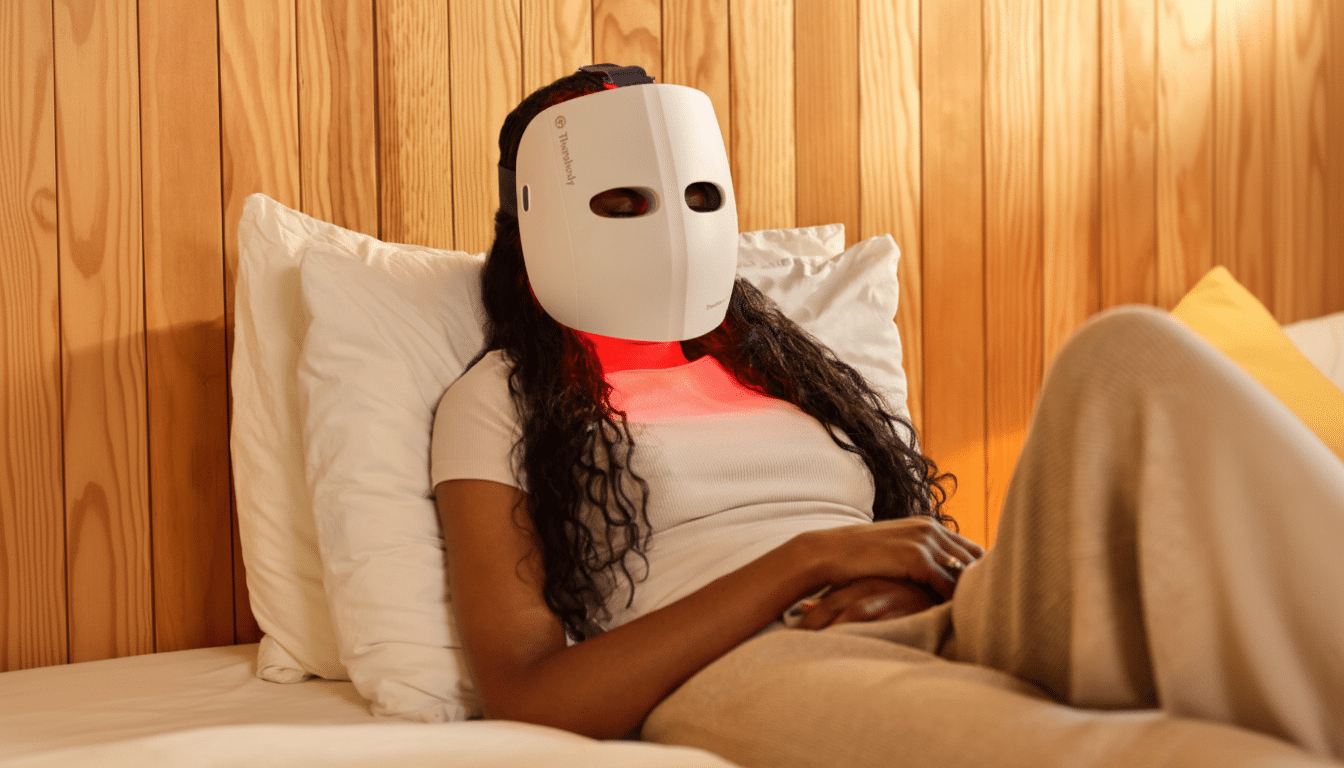 A person relaxing on a bed, wearing a white Therabody LED light therapy mask that emits a soft red glow around the chin area. Filename : therabody ledmask bed.png