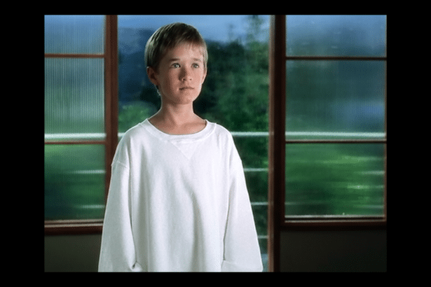 A young boy with short brown hair, wearing a white oversized long -sleeved shirt , stands thoughtfully in front of a large window with multiple panes looking out at a blurry green background .