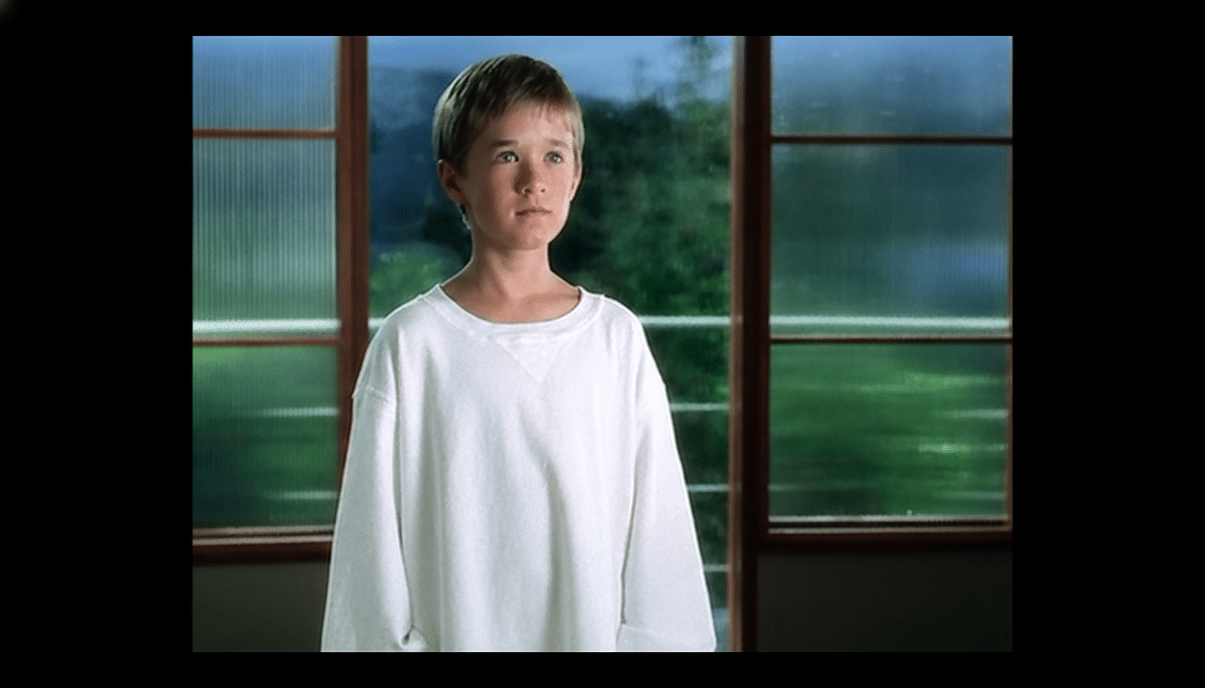 A young boy with short brown hair, wearing a white oversized long -sleeved shirt , stands thoughtfully in front of a large window with multiple panes looking out at a blurry green background .