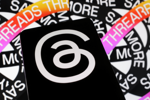 A close-up of a smartphone displaying the Threads app icon, which is a white looped ' a' on a black background. The phone is placed over a background of repeating THREADS text in white and black , with a colorful gradient banner partially visible at the top.