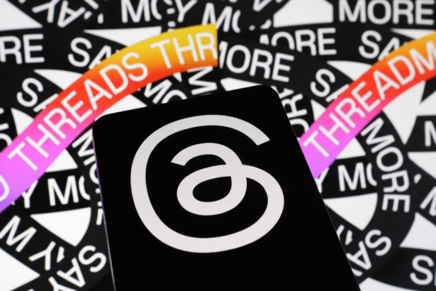 A black smartphone displaying the white Threads logo ( an a with a loop ) against a background patterned with THREADS text in black and white, with colorful gradient accents. Filename : threadslogo smartphone 1 69. png