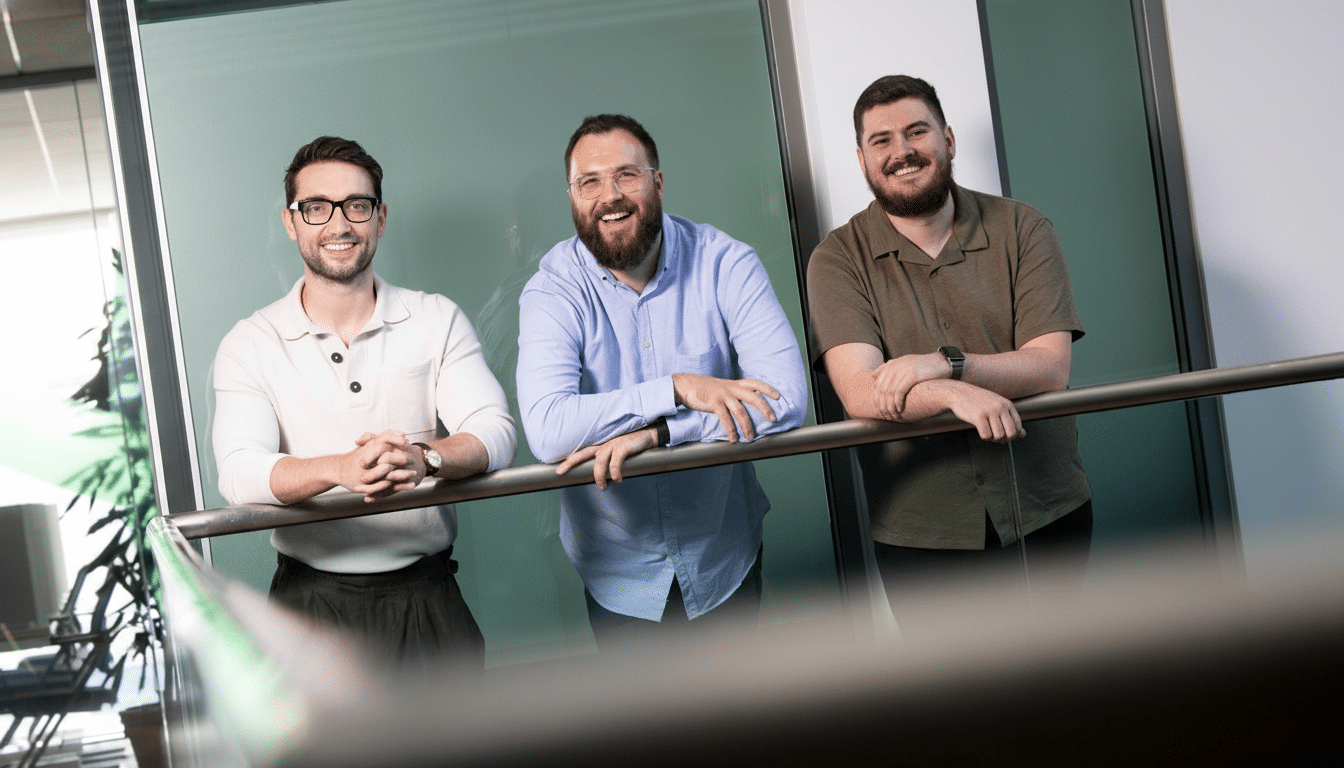 Three smiling men with be ards and glasses, leaning on a railing in a modern office environment .
