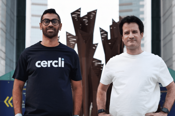 Two men , one wearing a black cercli t -shirt and glasses , and the other a white t-shirt , stand outside with a modern building and sculpture in the background.