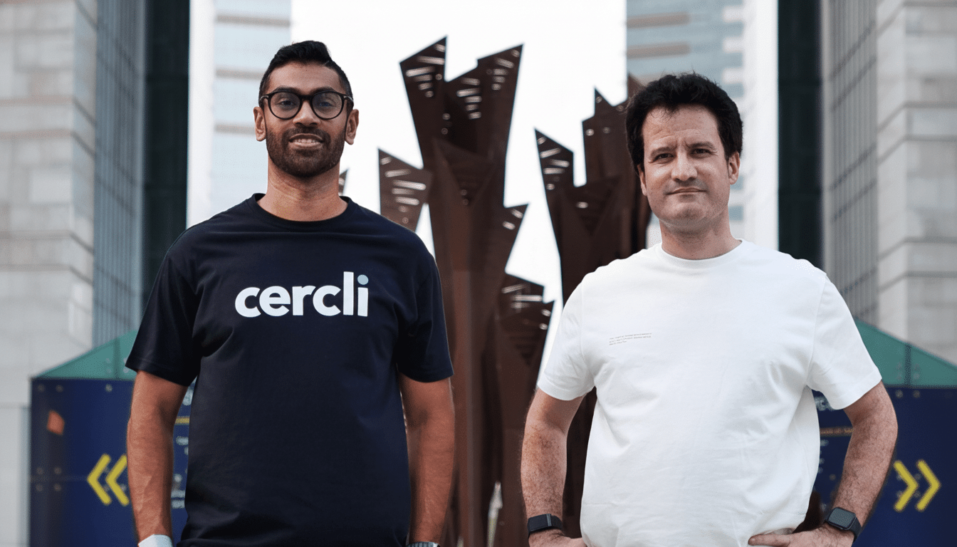 Two men , one wearing a black cercli t -shirt and glasses , and the other a white t-shirt , stand outside with a modern building and sculpture in the background.