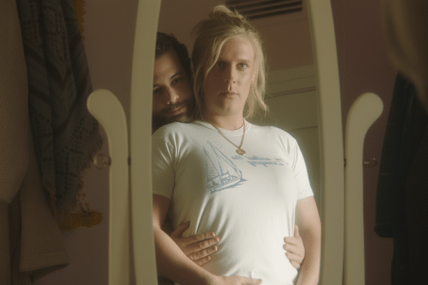 A man with blonde hair in a white t- shirt with a sailboat design, wearing a necklace with a gold pendant, looks into a mirror. Another man with dark