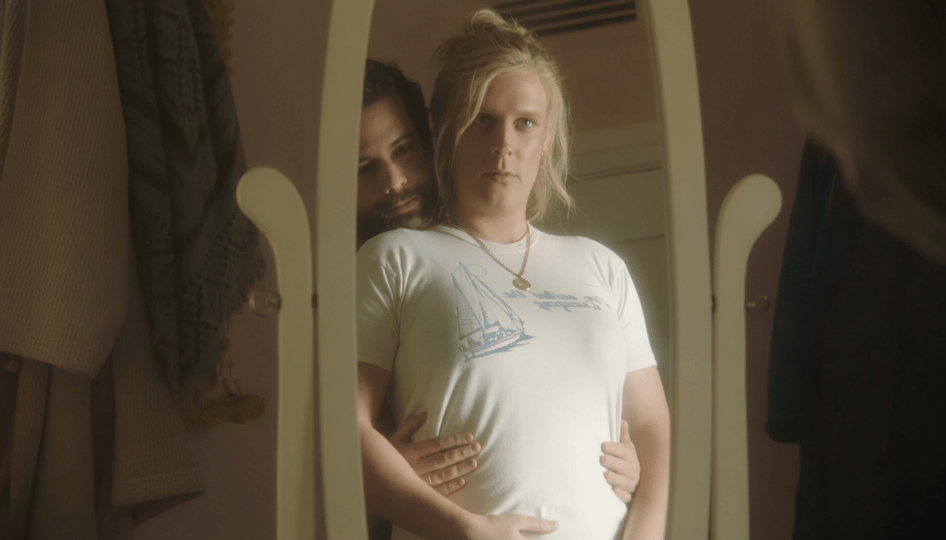 A man with blonde hair in a white t- shirt with a sailboat design, wearing a gold necklace, looks directly into a mirror. Behind him, another man with