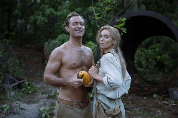 A man and a woman stand in a natural, outdoor setting. The man is shirtless and holding a yellow fruit, while the woman is wearing light - colored clo
