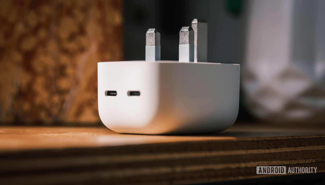 A white, rectangular charging adapter with two USB -C ports and two metal pr ongs for a UK socket, resting on a wooden surface.