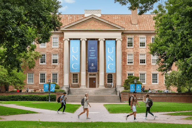 The main building of the University of North Carolina at Chapel Hill, a large brick structure with columns and blue banners, with students walking on pathways in front of it.