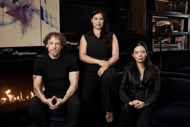 Three individuals , two women and one man, posing in a professional setting with a fireplace and modern artwork in the background.