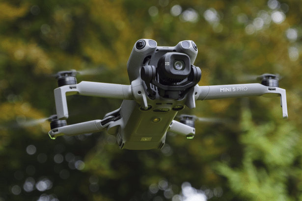A DJI Mini 5 Pro drone hovering in front of a blurred green foliage background, resized to a 16: 9 aspect ratio.