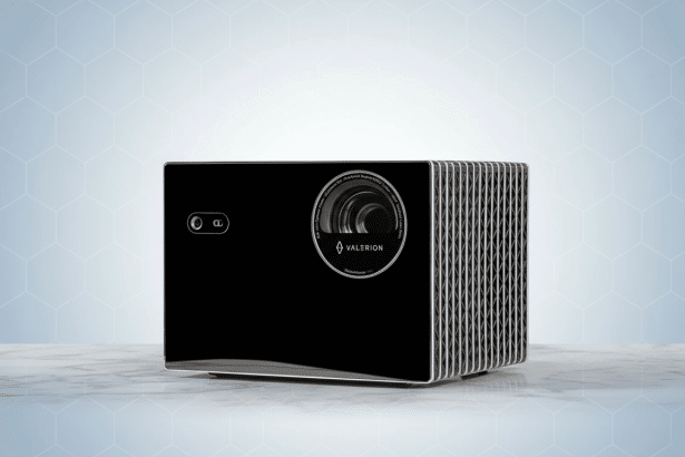 A sleek , black Valer ion projector with a silver ribbed side, sitting on a white marble surface, against a professional light blue background with a subtle hexagonal pattern.